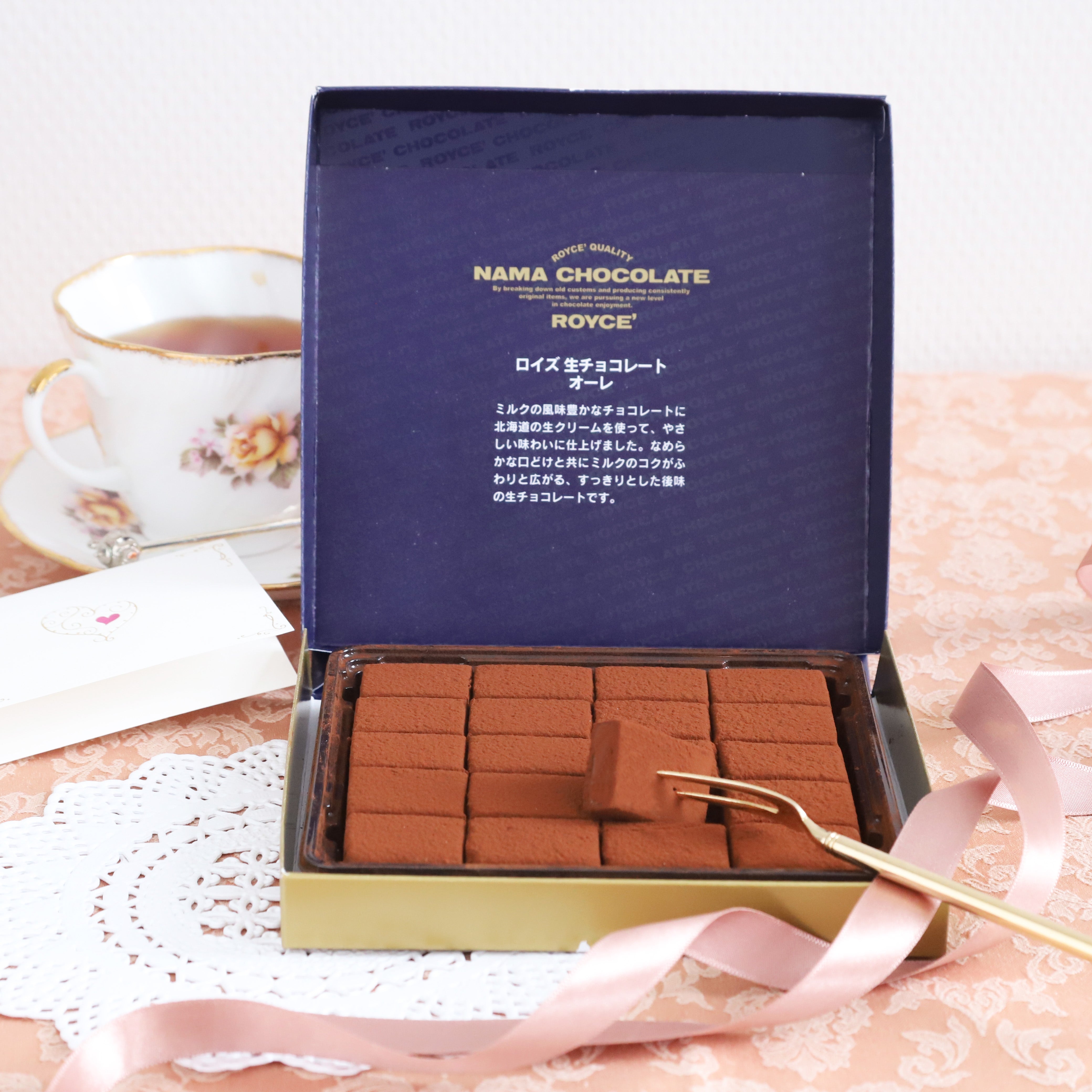 Open box of Royce “Nama Chocolate” on a gold-trimmed tray, with a single Nama on a fork. A teacup and greeting card in the background.