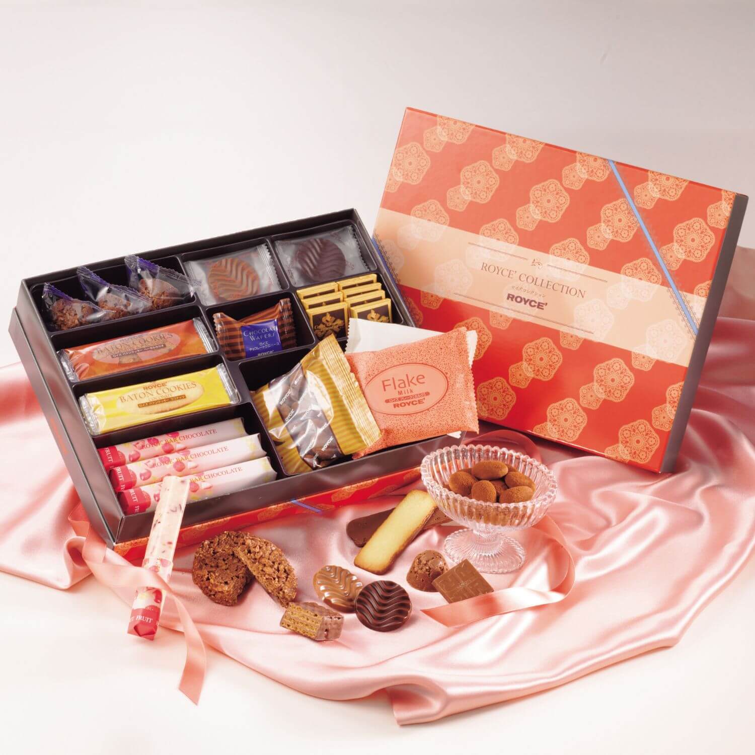 Open gift box of assorted Royce’ sweets featuring cookies, chocolates, and truffles, with lid labeled “Royce Collection,” arranged on satin backdrop.