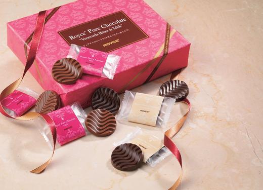 Gift box labeled Royce’ Pure Chocolate with wrapped chocolate coins in pink/red and cream, unwrapped discs showing wavy surfaces, and decorative ribbons.