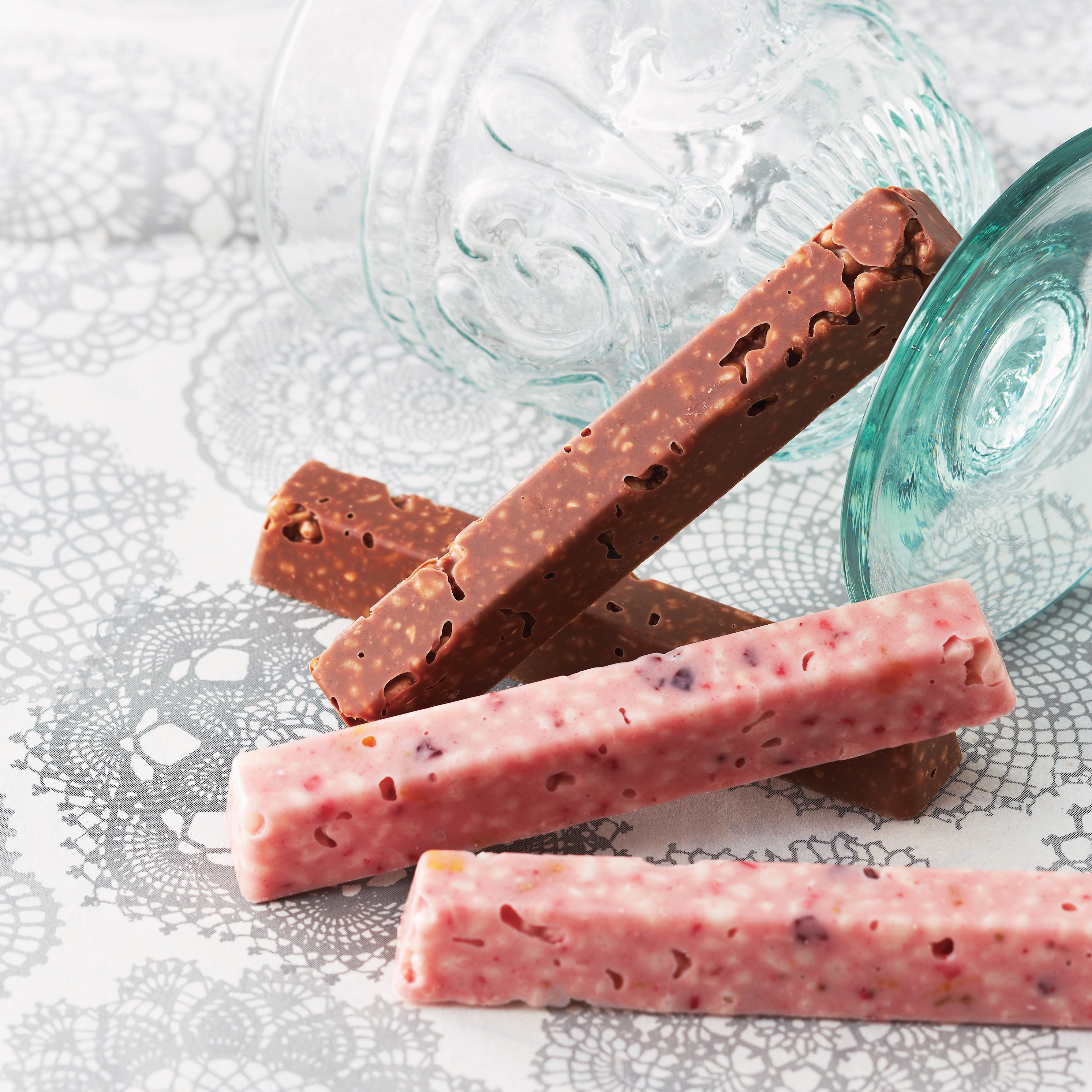 Stack of candy bars with milk and pink chocolate coatings, featuring crisped rice bits, on a lace-patterned surface with glass vessels in the background.