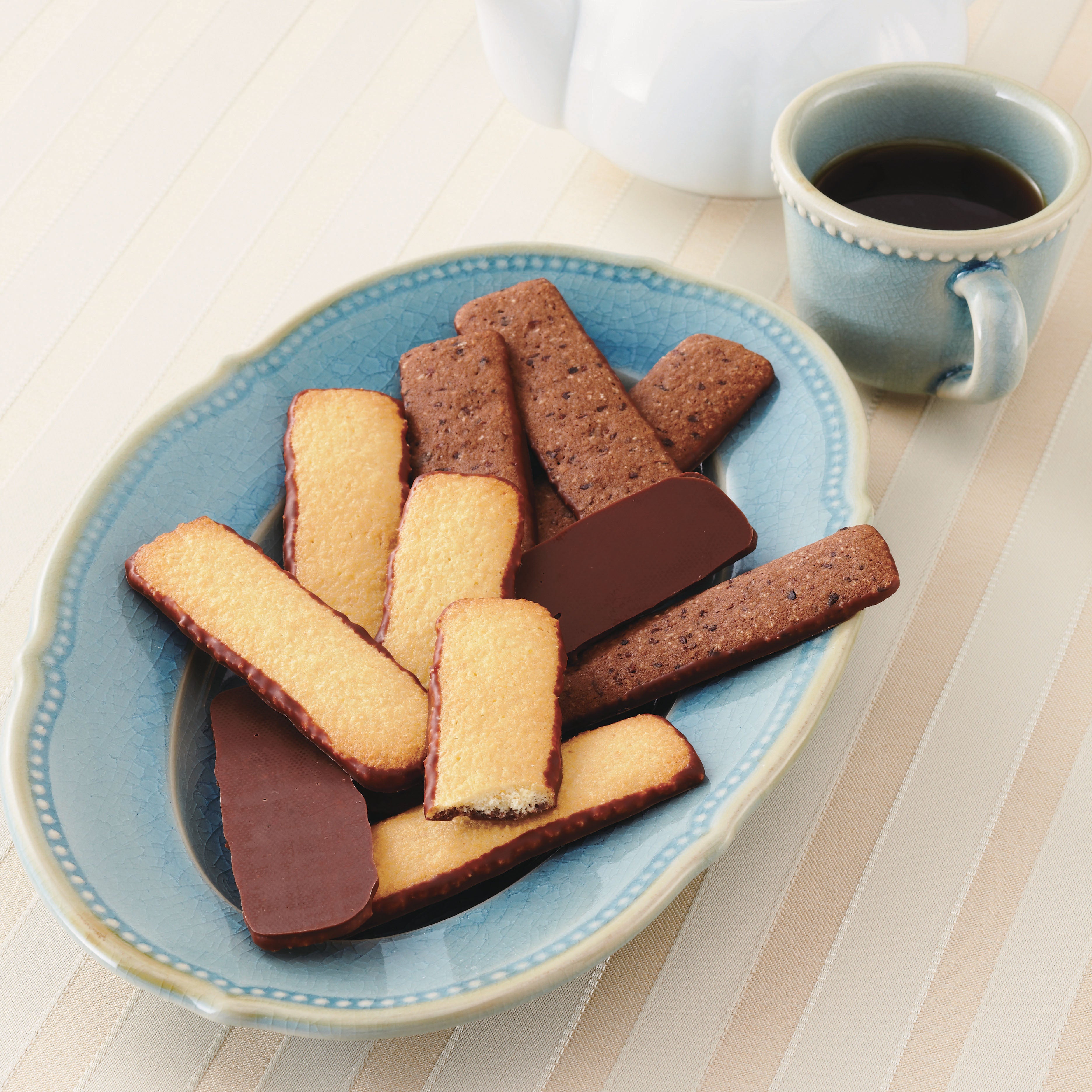 Blue ceramic dish with chocolate-dipped shortbread and chocolate cookies, accompanied by a matching mug of black coffee and a teapot on a striped tablecloth.