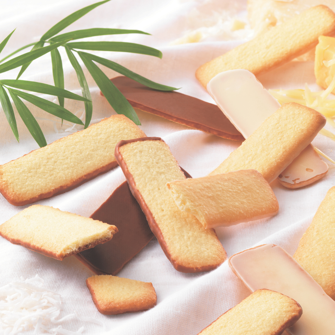 Assorted rectangular butter cookies, some half-dipped in milk or white chocolate, garnished with green leaves and shredded coconut on a light cloth.
