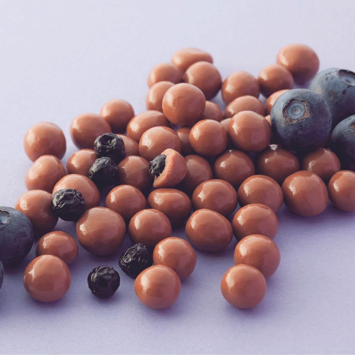 Round, glossy light-brown spheres, likely chocolate-covered blueberries, mixed with dark dried berries and fresh blueberries. A blend of coated and natural fruit.