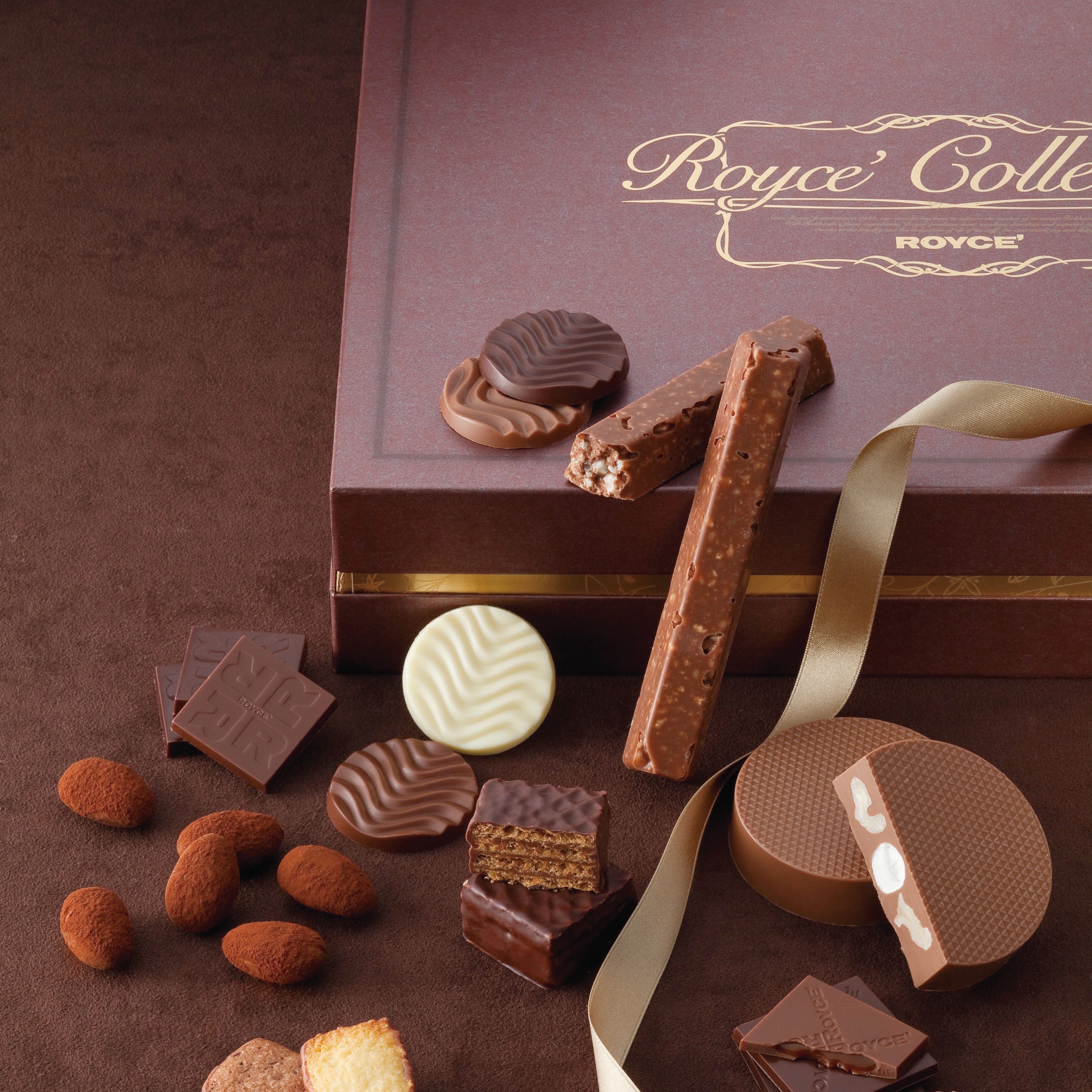 Luxury Royce’ chocolate gift set with assorted confections: wavy discs, nut-studded sticks, imprinted bars, cocoa almonds, and marshmallow-filled tin adorned with a gold ribbon.