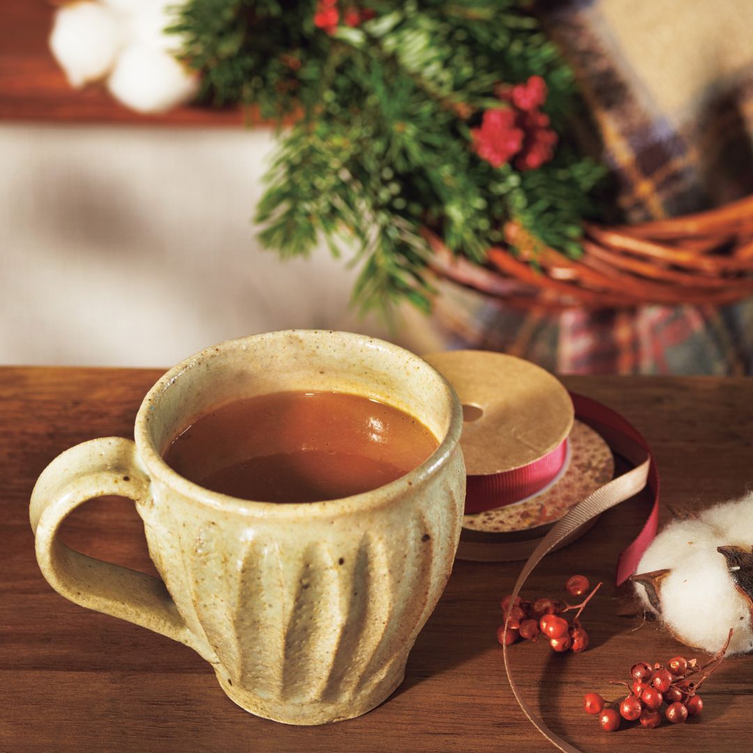 Cozy holiday scene with a ceramic mug on a wooden table, evergreen sprigs, cotton bolls, red berries, and spools of ribbon.