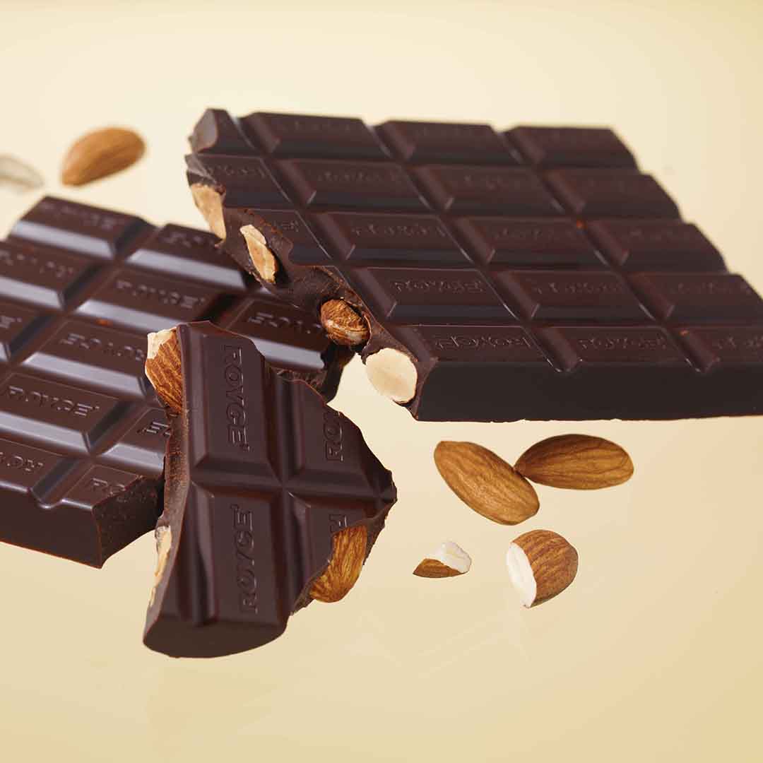 Dark chocolate squares with whole almonds visible inside; broken pieces and scattered almonds illustrate the rich, nutty texture and flavor.