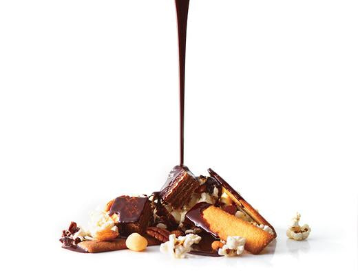 A small pile of chocolate bars, butter cookies, popcorn, nuts, and caramel nuggets drenched in rich dark chocolate sauce on a white surface.