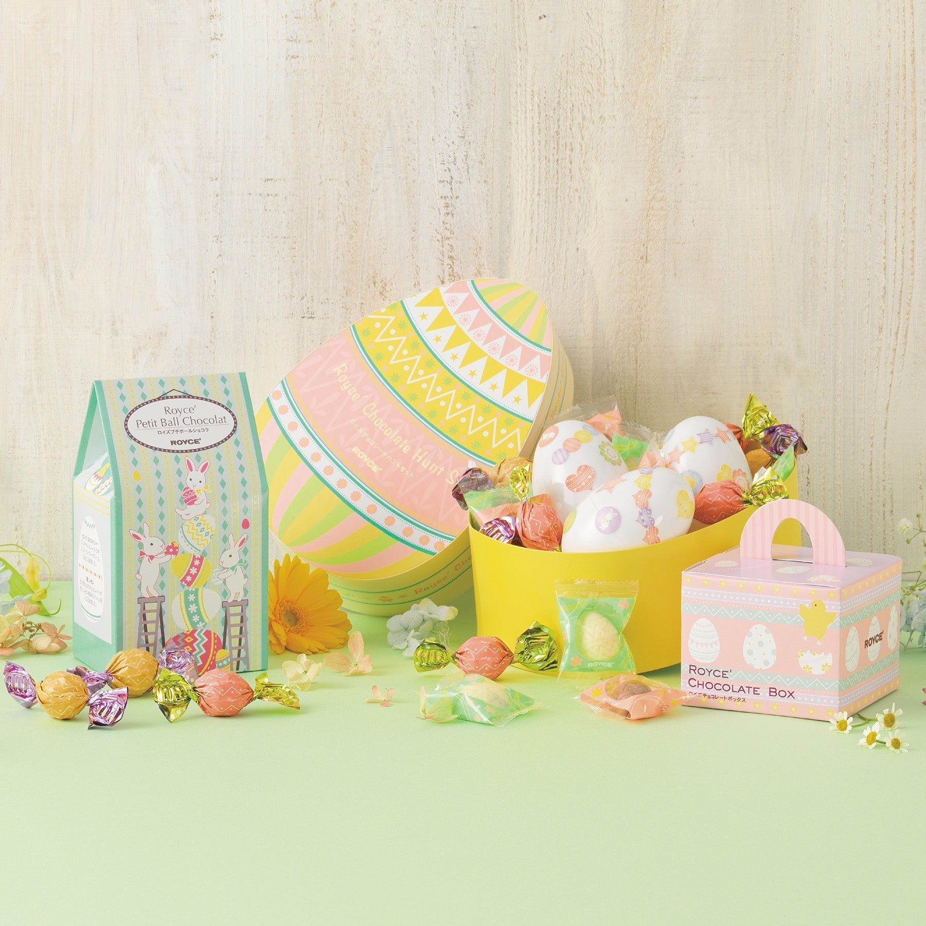 Pastel Easter display featuring a striped egg-shaped gift box, Royce’ chocolate boxes, a basket of chocolate eggs, bonbons, and scattered spring flowers.