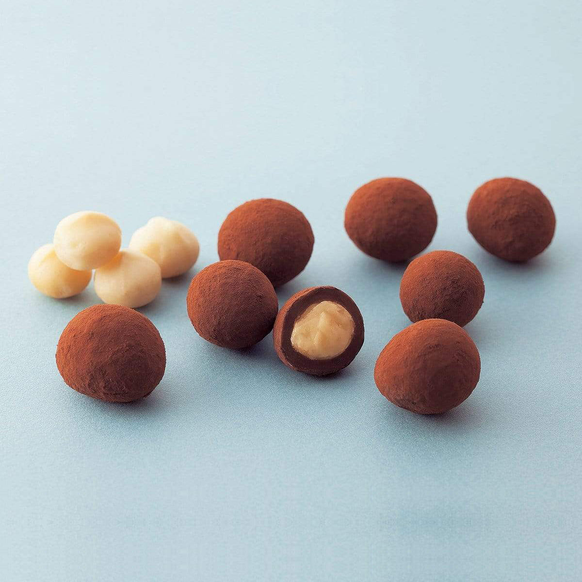 Round chocolate truffles, dusted with cocoa, displayed with a cut piece revealing creamy interior, alongside a few white-coated confections, likely white chocolate or nut centers.