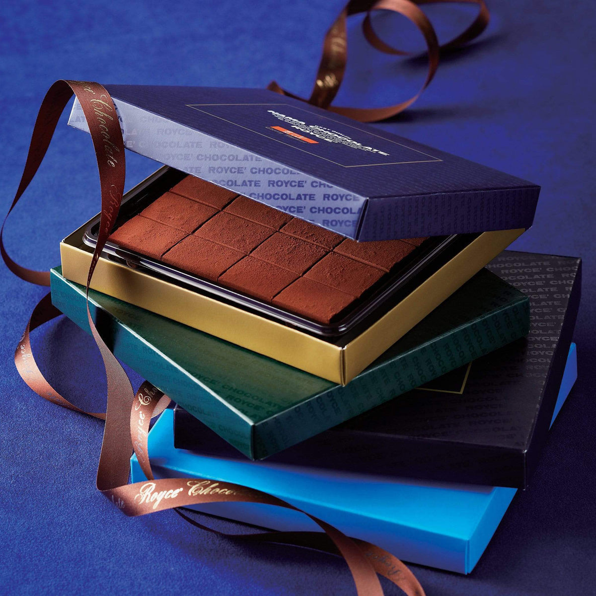 A stack of blue, green, and navy gift boxes tied with a Royce’ Chocolate ribbon, revealing cocoa-dusted Nama chocolate truffles inside.