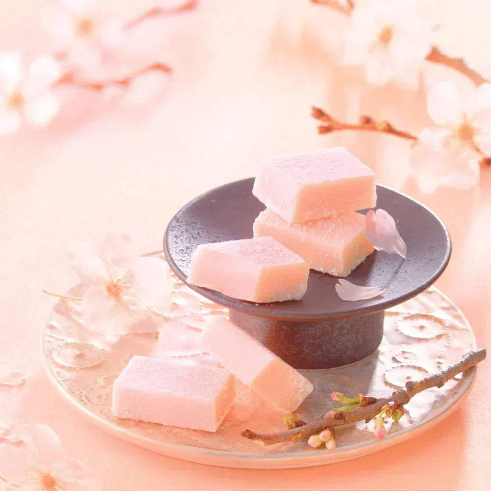 Pedestal dish with pastel-pink, sugar-coated Namas on a dark tray, adorned with cherry-blossom petals and a twig for decoration.