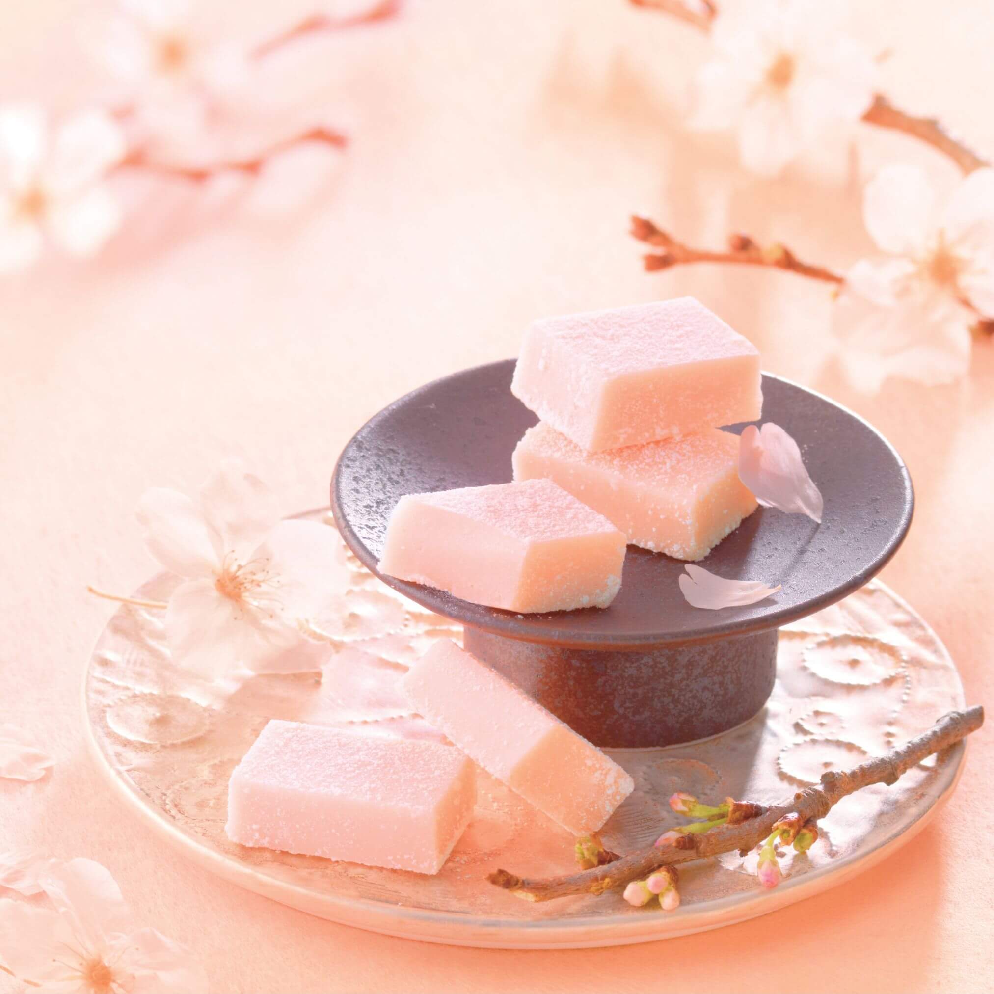 A dark-toned serving stand with pale-pink sakura-flavored Namas, surrounded by cherry-blossom flowers and twigs, creating a soft, springtime ambiance.