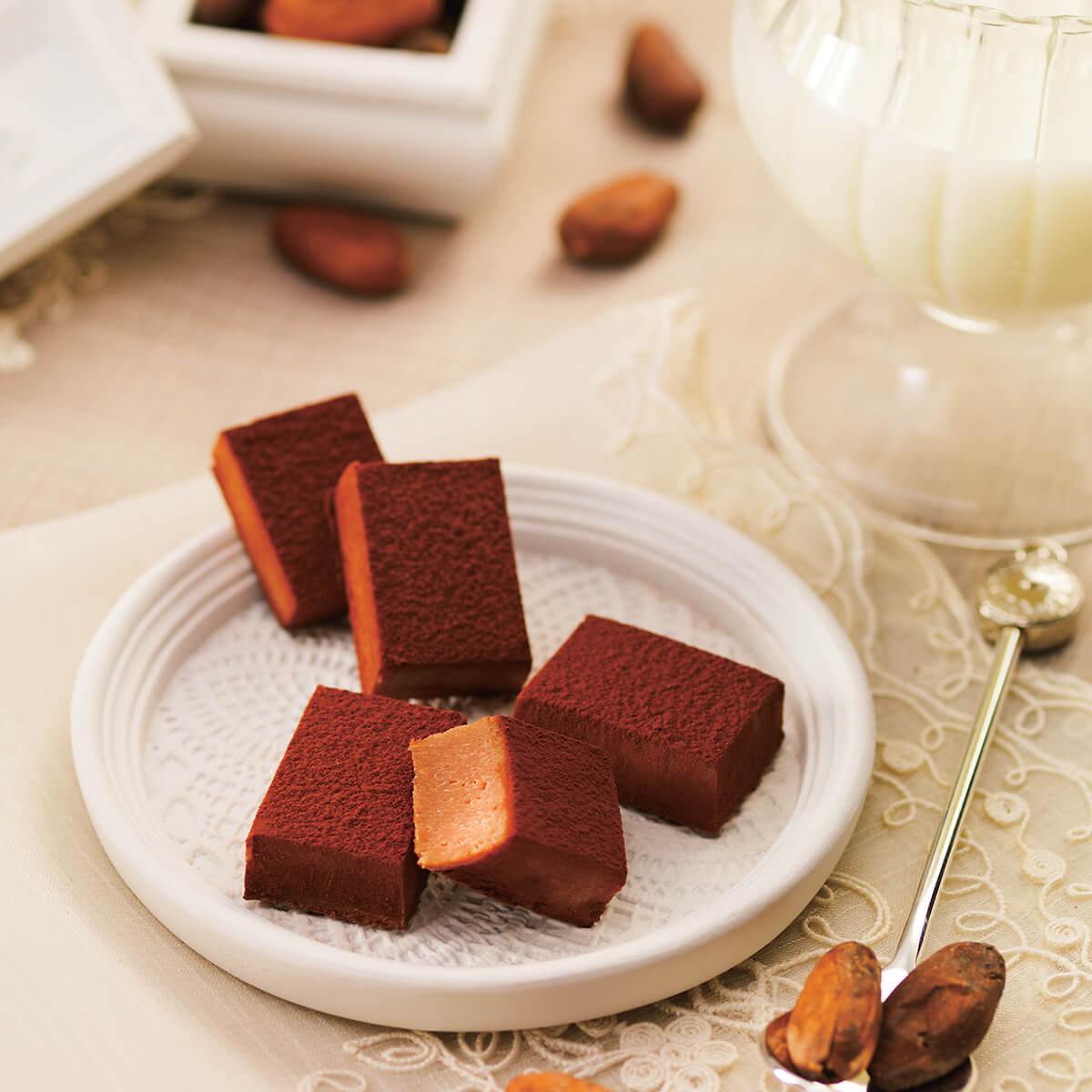 Stack of cocoa-dusted chocolate Namas on a white plate with a gold spoon and cocoa beans nearby; glass of milk in background.