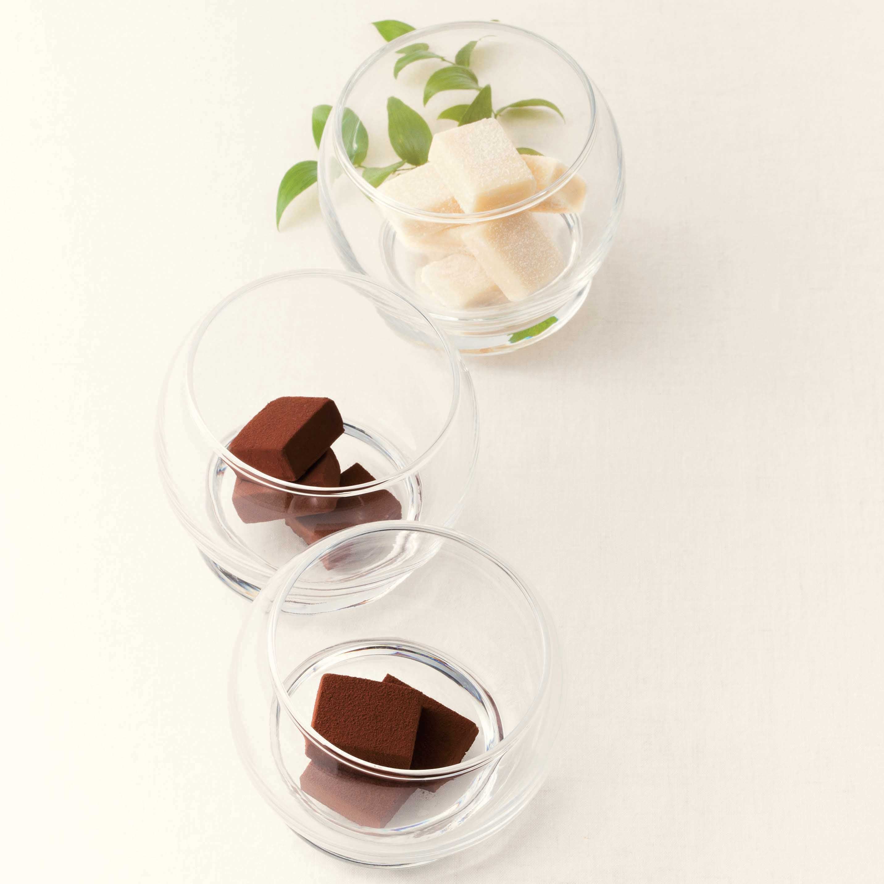 Stack of clear glass bowls with white sugar-coated Namas and cocoa-dusted chocolate Namas, garnished with green leaves, on a neutral background.