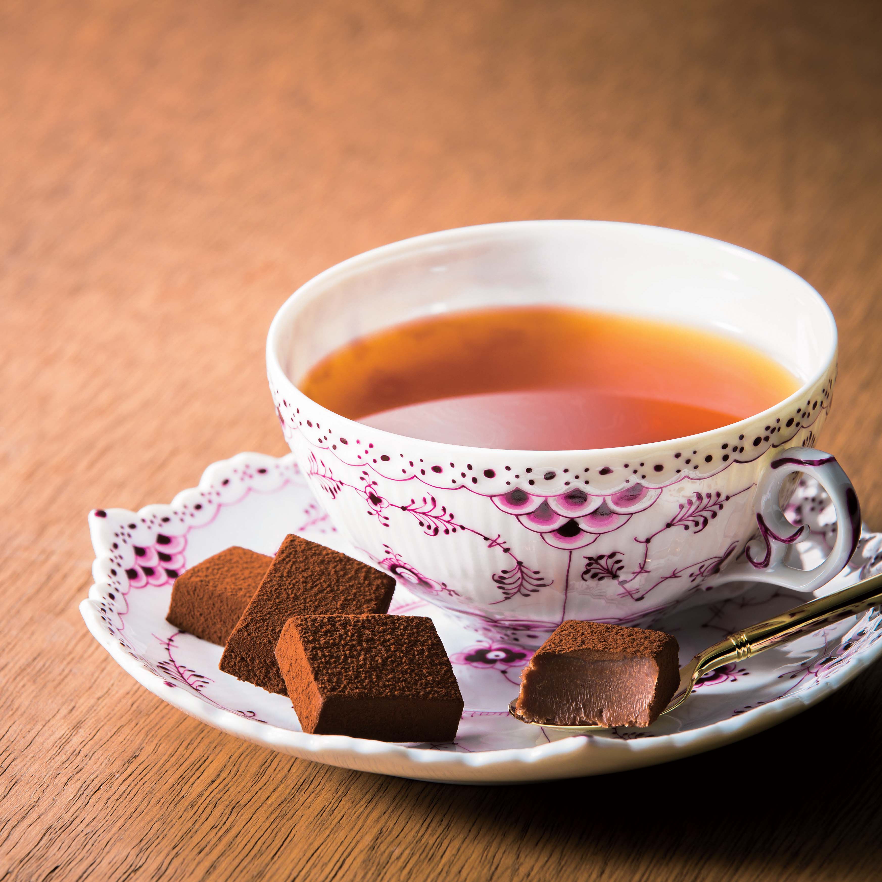 Porcelain teacup with floral pattern on saucer, filled with tea, surrounded by cocoa-dusted chocolate Namas on wooden tabletop.