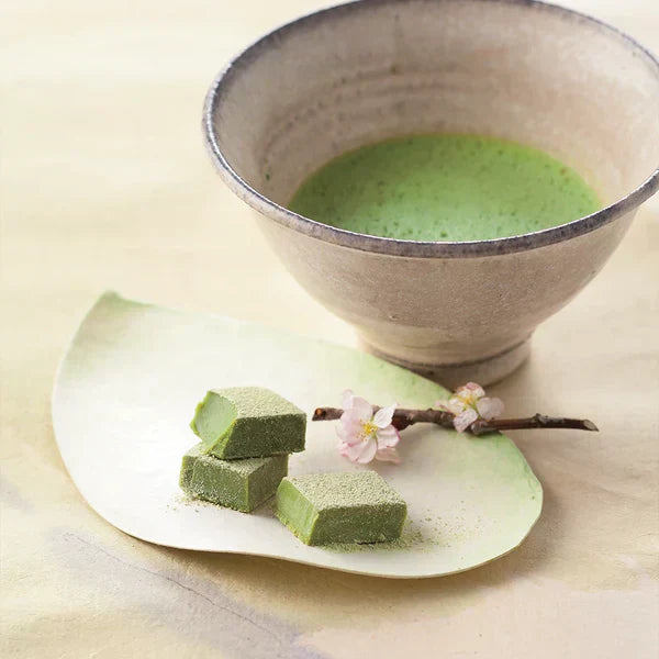 Japanese-style tea setup with a ceramic chawan of frothy matcha, leaf-shaped plate holding matcha Namas, and a sakura twig accent.