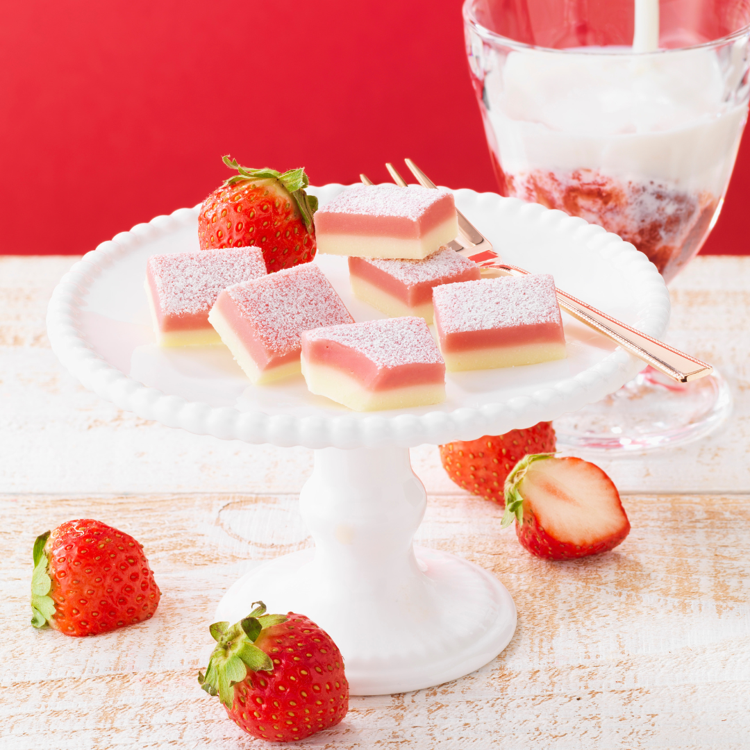 A white cake stand with pink and ivory Namas, surrounded by fresh strawberries, a gold fork, and a glass of strawberry-swirled yogurt.