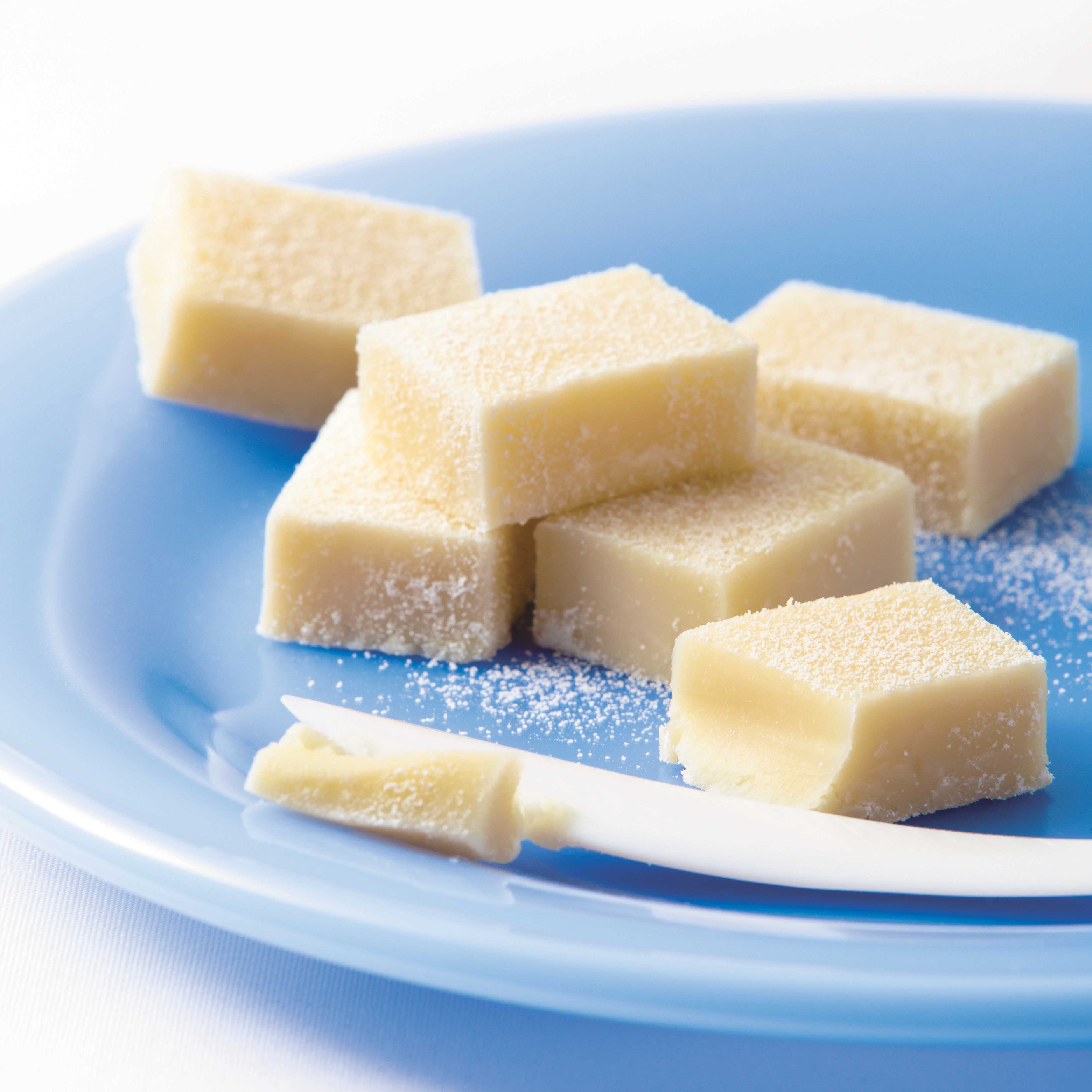 Close-up of small, ivory-colored Namas resembling white chocolate or vanilla fudge, lightly dusted with sugar, arranged on a glossy blue plate.