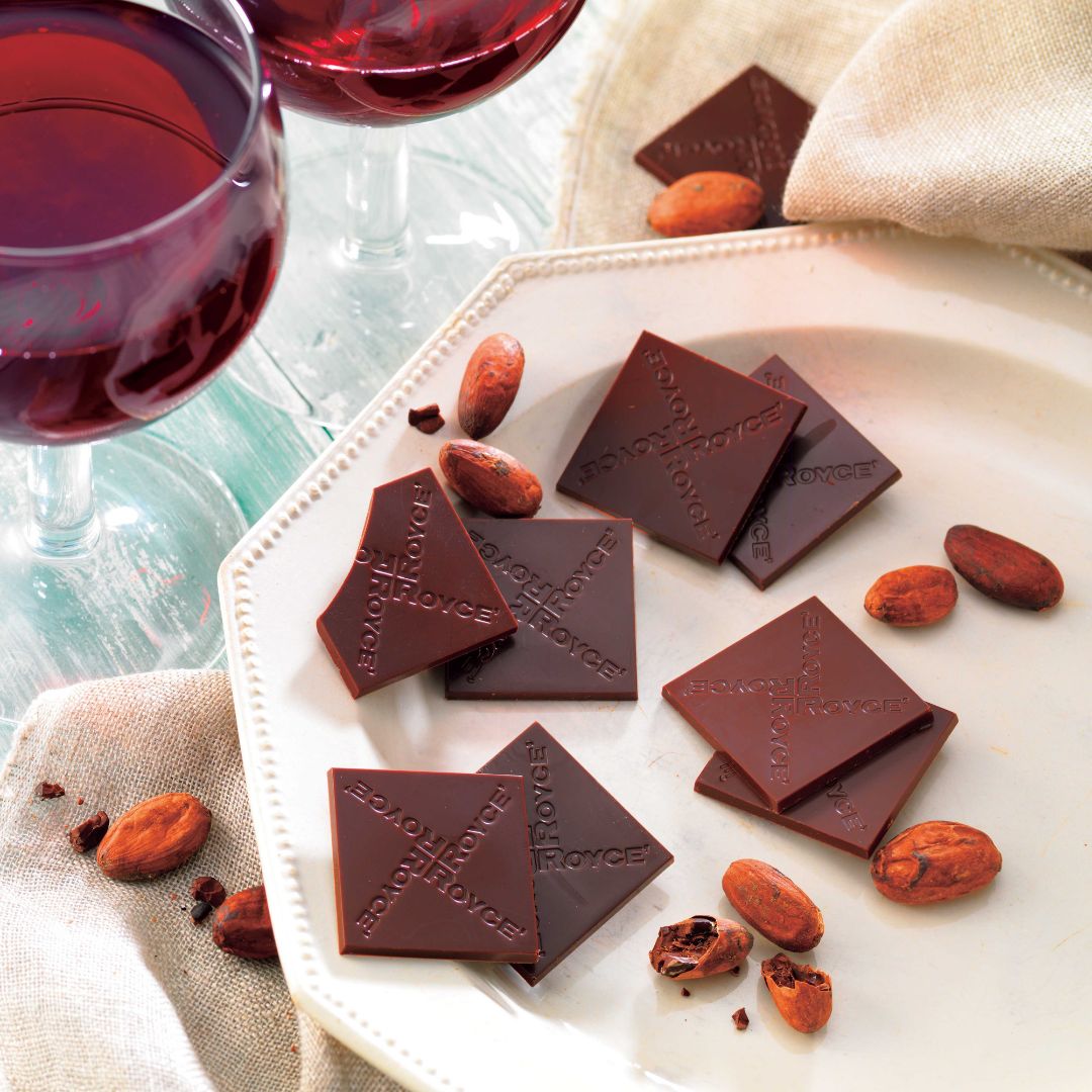 Dark-chocolate squares marked ROYCE on a white plate with cacao beans, accompanied by two red wine glasses on a linen napkin.