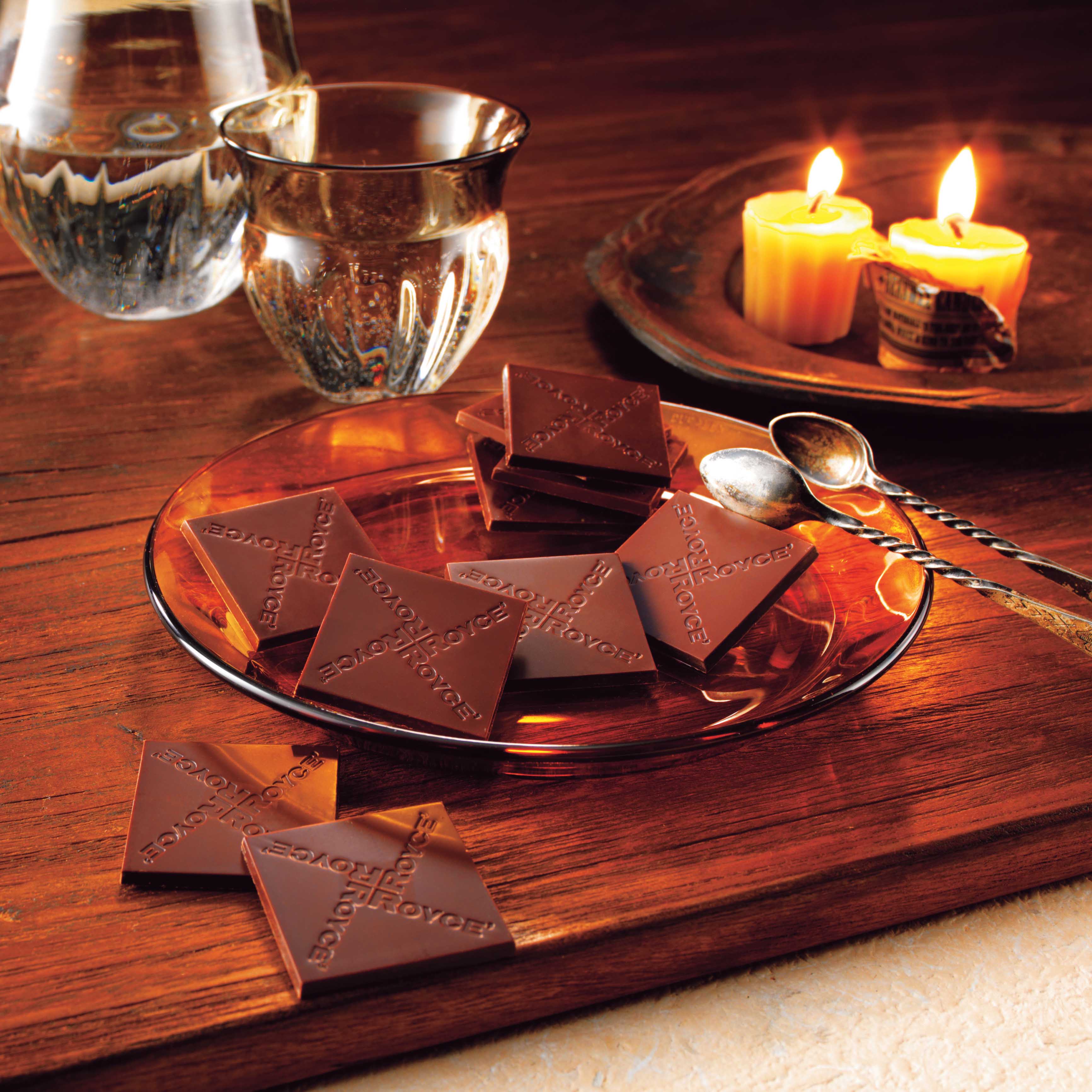 Cozy tabletop with ROYCE chocolate on an amber plate, twisted-handle spoons, a glass carafe, tumbler, and lit candles casting a warm glow.