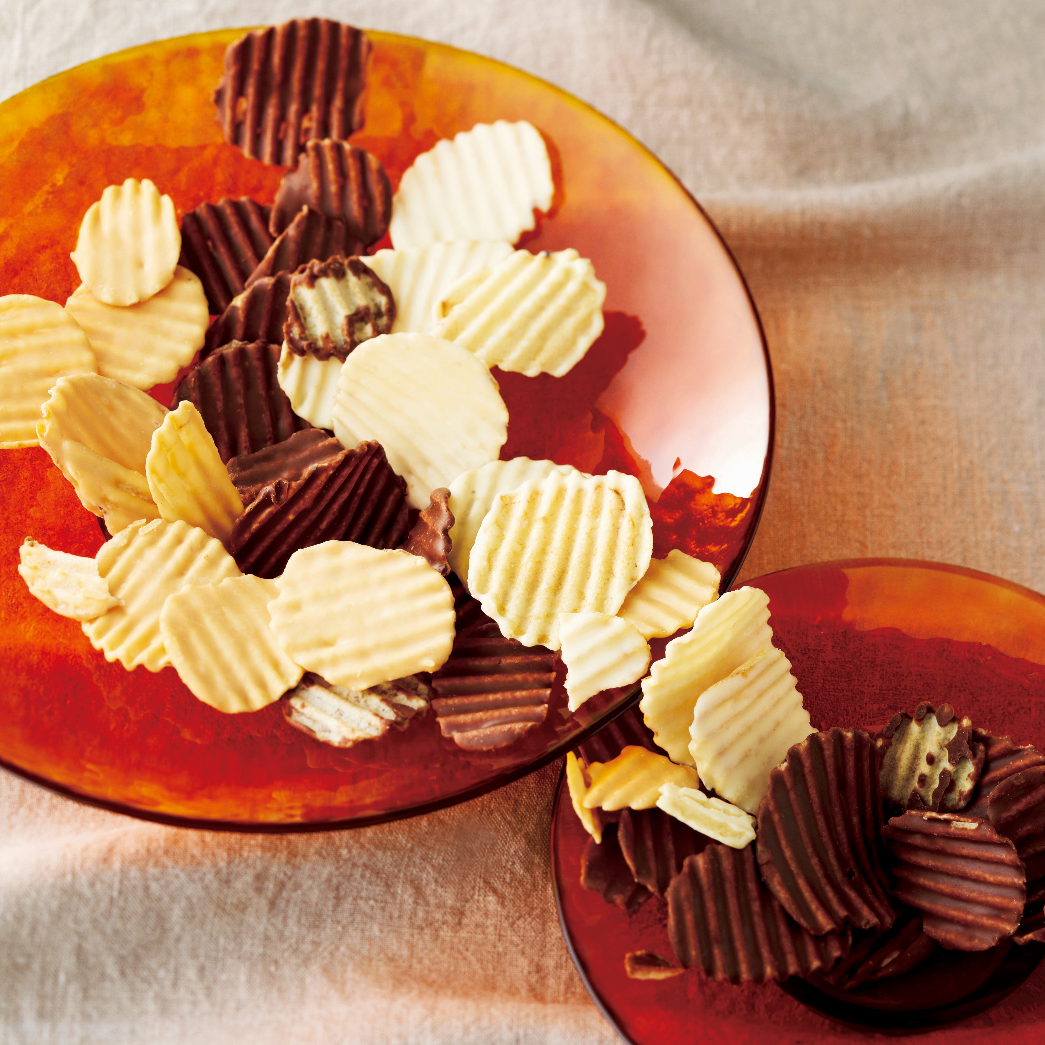 Close-up of wavy potato chips half-dipped in white and milk/dark chocolate, arranged on translucent orange plates, highlighting their texture and chocolate coating.