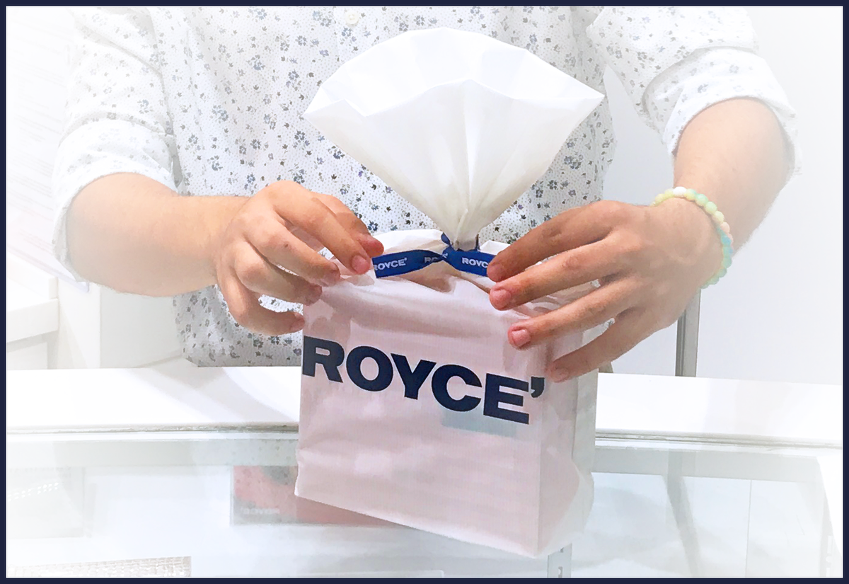 Hands tying a blue ribbon on a Royce' gift bag, preparing a chocolate package in a shop setting.