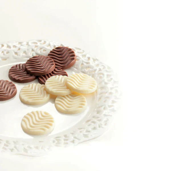 A decorative white plate holds round chocolate discs, featuring milk and white chocolate varieties, each with a delicate wavy-line pattern on top.
