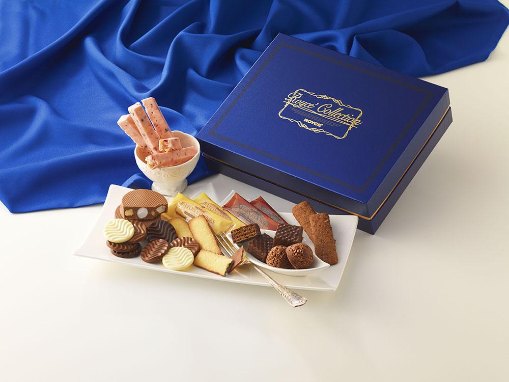 Gift-style confection set in a deep blue box with assorted chocolates, cookies, truffles, and strawberry-coated wafer sticks arranged on a white platter.
