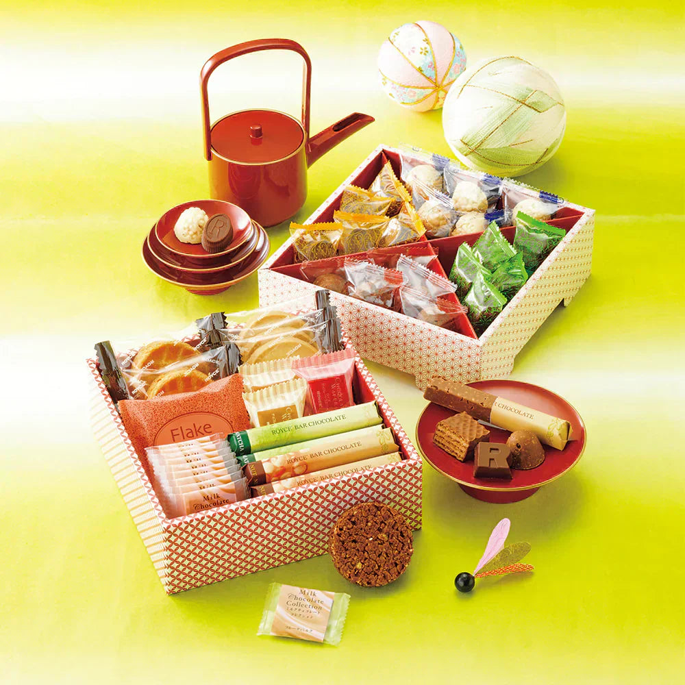 Gift set with patterned trays of chocolates, cookies, wafer bars, red lacquer dishes, a matching teapot, and decorative temari balls in the background.