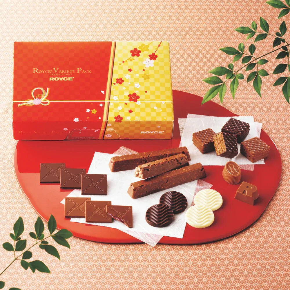 Assorted Royce chocolates on a lacquer-red tray, featuring variety pack box, embossed squares, wafer cubes, nut bars, discs, and molded pieces with decorative leaves.