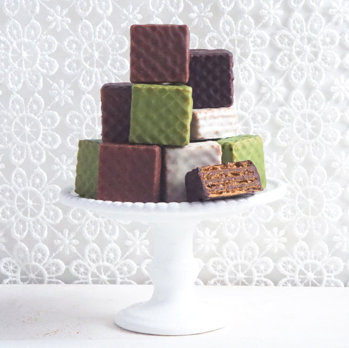 A white pedestal cake stand holds assorted chocolate-coated wafer cubes, including milk, dark, white, and matcha flavors, one cut open showing layered wafers.