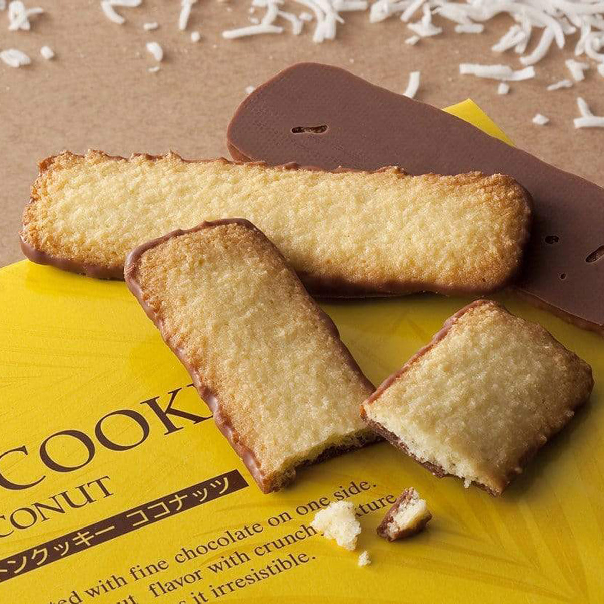 Close-up of rectangular coconut cookies, one side dipped in chocolate, on yellow packaging with scattered coconut shreds; one cookie is broken, showing its interior.