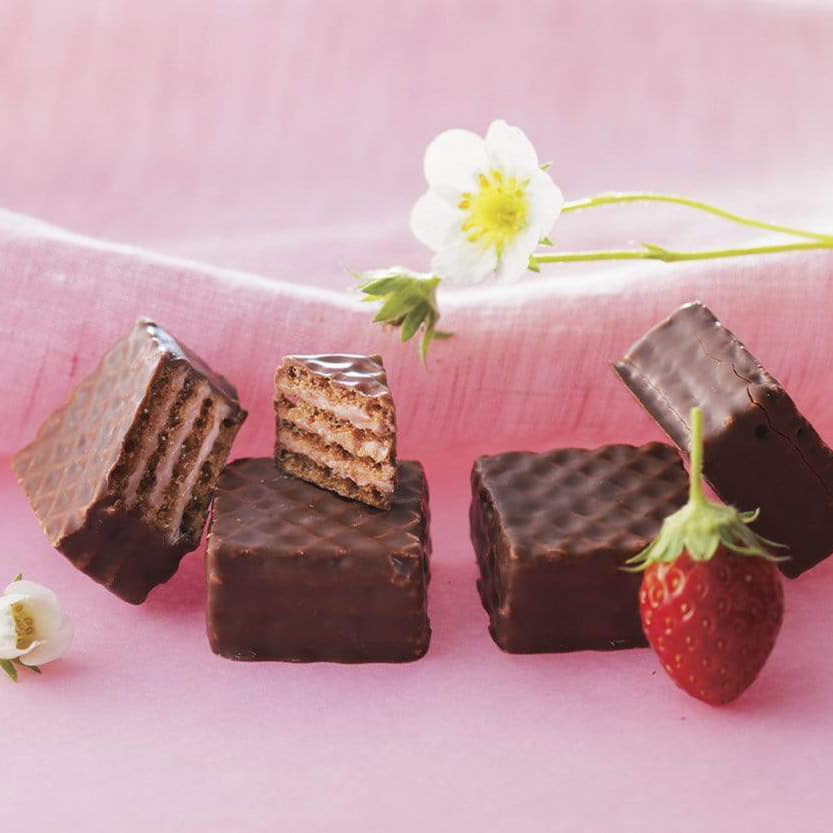 Chocolate-coated wafer squares with visible layers of wafer and cream, accompanied by a fresh strawberry and a white strawberry blossom.