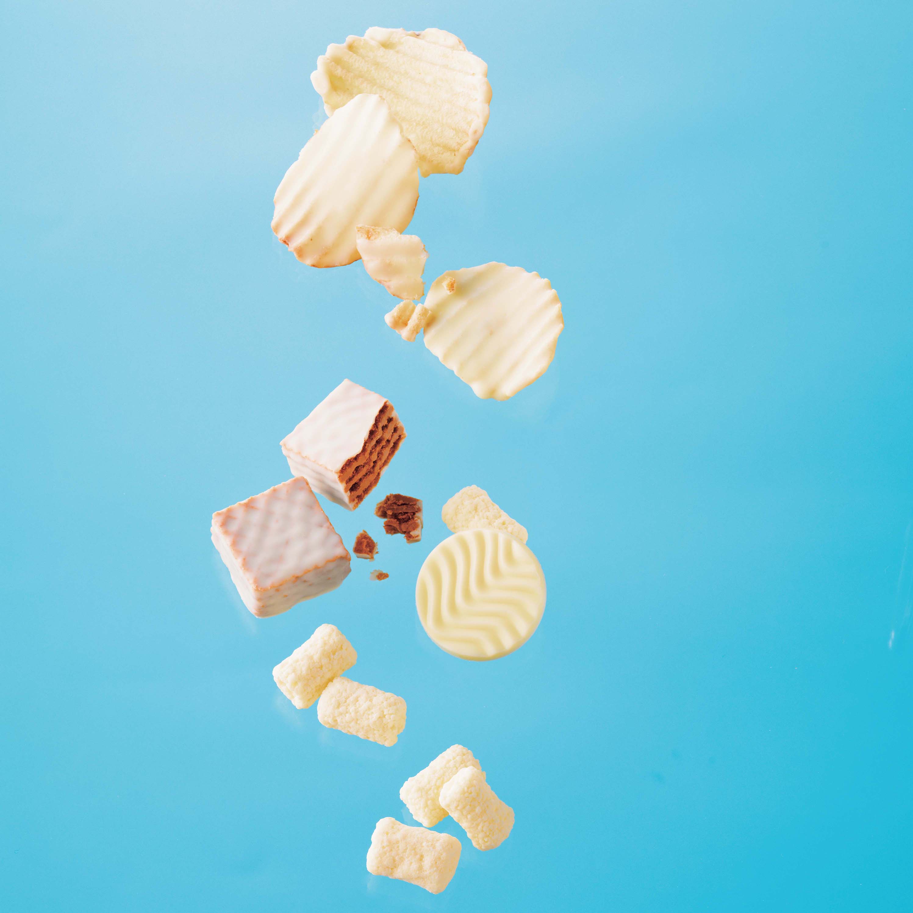 An array of pale-colored snacks, including white-chocolate-coated wafers and crinkle cookies, appear suspended mid-air with puffed corn pieces.