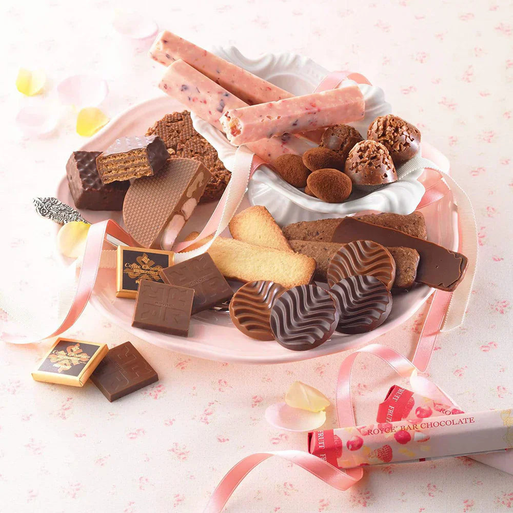 Assorted confections on a dessert plate: chocolate-coated wafers, truffles, butter cookies, fruit-studded chocolate sticks, and wrapped squares, adorned with ribbons and rose petals.
