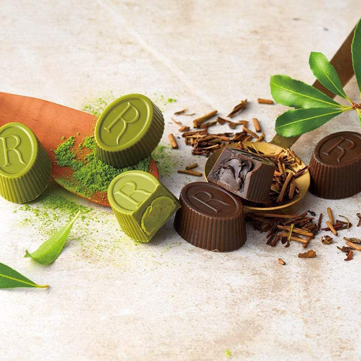 Stylized image of matcha and cocoa/tea-flavored chocolate bonbons embossed with R, arranged with a wooden spoon, matcha powder, tea leaves, and fresh green leaves.