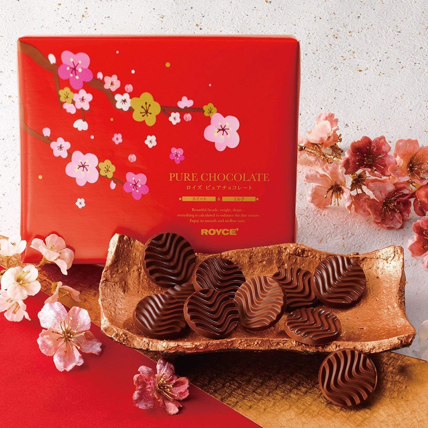 Decorative red box of ROYCE’s PURE CHOCOLATE with cherry-blossom motifs, metallic-gold platter with ripple-patterned chocolate discs, surrounded by ornamental cherry blossoms.