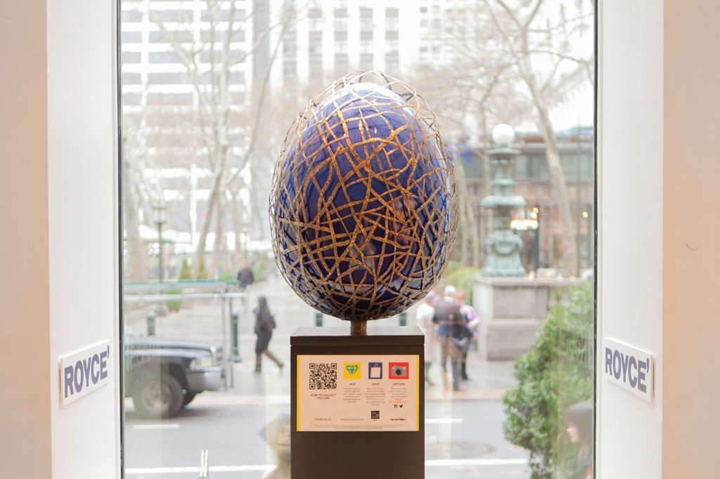 Cobalt blue Faberge egg sculpture with gold crack pattern on black pedestal in Royce' storefront, featuring a QR code and icons. City sidewalk visible outside.