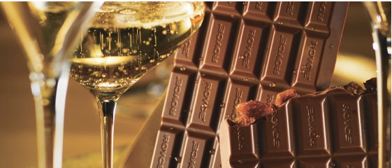Bars of milk chocolate, some with nuts, arranged beside two glasses of pale bubbly beverage, likely white wine or champagne.