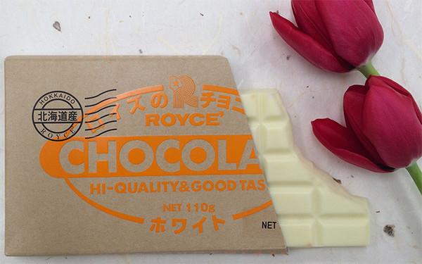 Partially unwrapped Royce white chocolate bar with HI-QUALITY & GOOD TASTE text and Hokkaido origin, next to two red tulip blossoms.