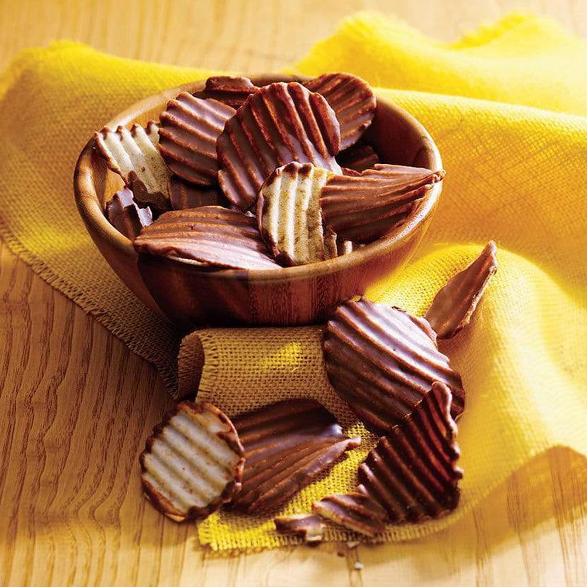 Wooden bowl filled with ridged potato chips partially dipped in milk chocolate, with several chips spilled onto a yellow cloth-covered table.