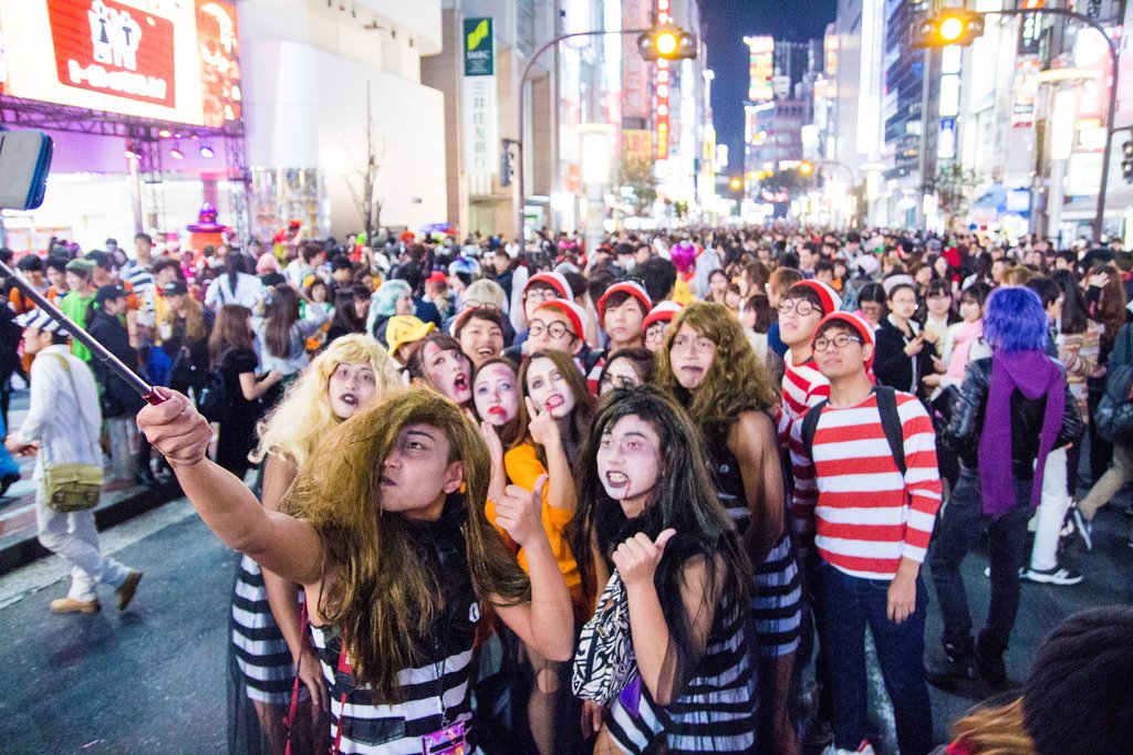 A crowded Tokyo street scene with costumed people, including Where's Waldo? characters and zombies, posing for a selfie amidst bright city lights.
