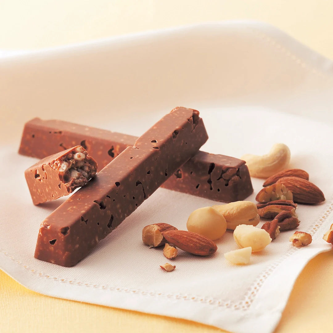 Stack of milk-chocolate nut bars with a puffed-rice interior, surrounded by assorted whole and halved nuts including almonds, cashews, macadamias, and pecans.