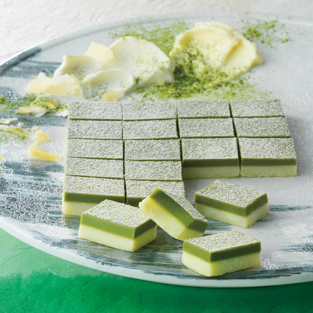 Platter of two-layered confections with white-chocolate base and matcha top, dusted with matcha powder, surrounded by white-chocolate shavings and extra matcha powder.