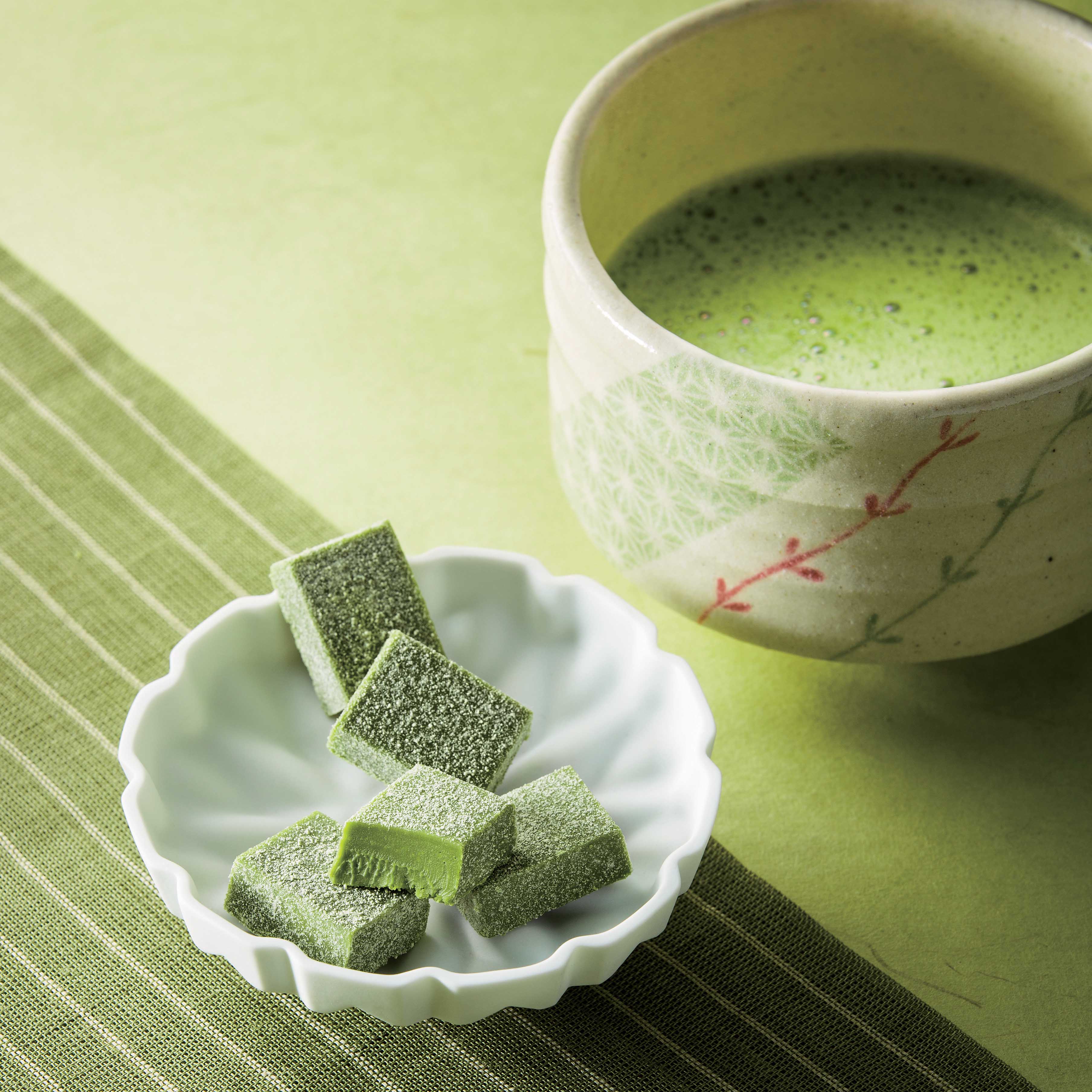 Scalloped dish with green tea-flavored chocolate squares in foreground; ceramic cup of frothy matcha tea in background on green linens.