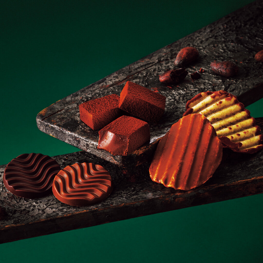 Assorted chocolate confections on dark slab: ripple-patterned disks, cocoa-dusted truffles, red-tinged chocolate chips, and scattered cocoa beans.