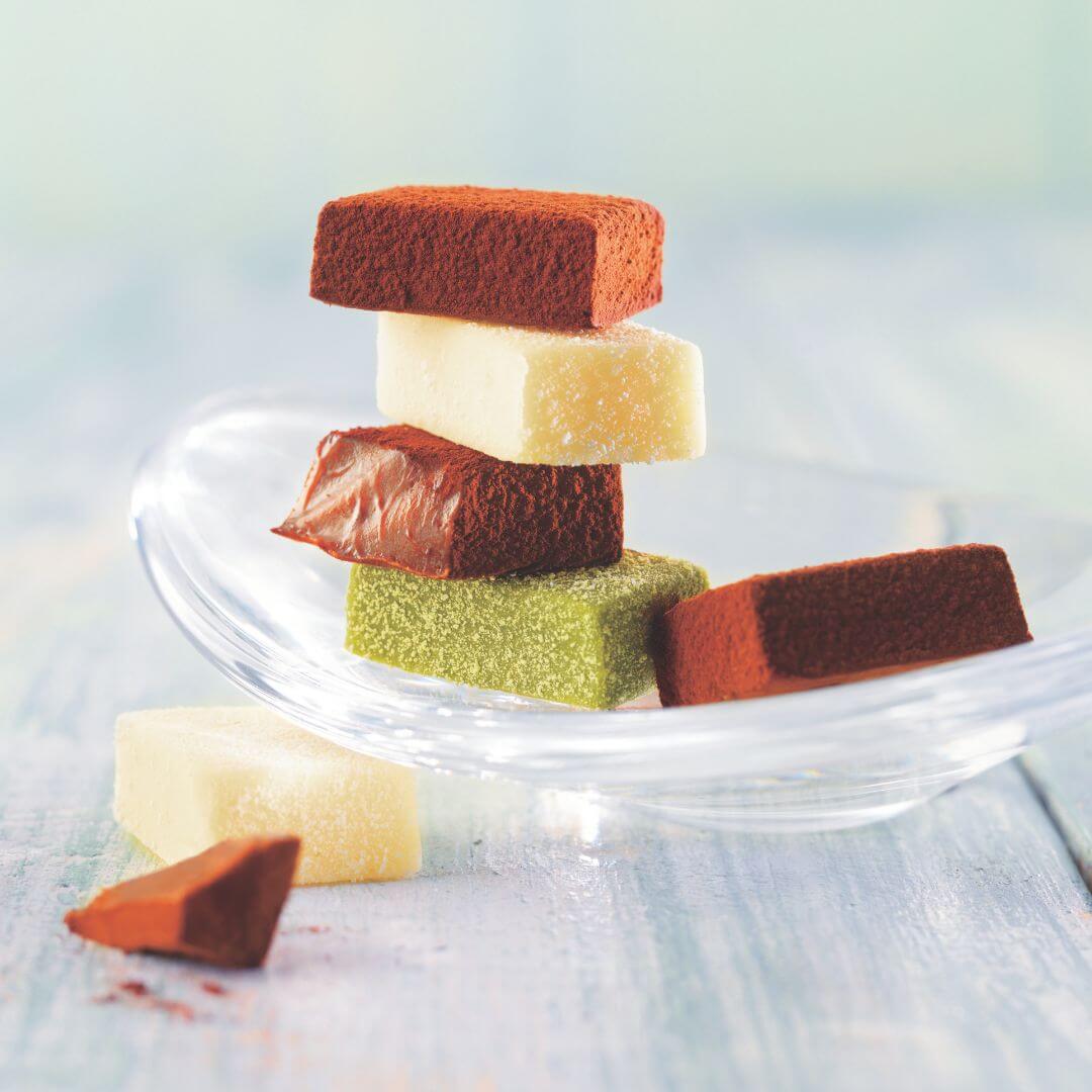 Stack of chocolate Namas: cocoa-dusted, vanilla-dusted, glossy chocolate, matcha-dusted, and another cocoa-dusted, elegantly arranged on a curved glass dish.