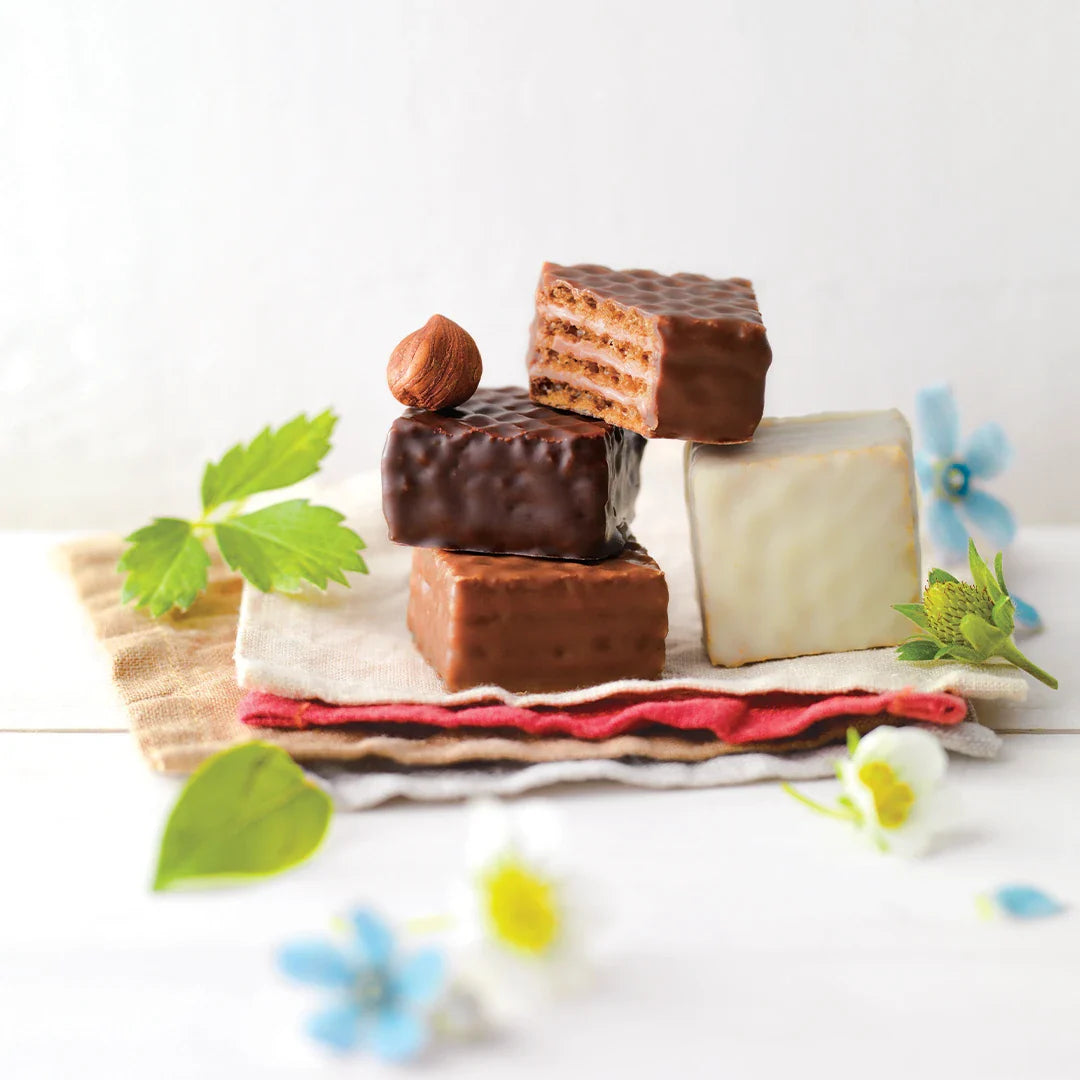 Stack of iced wafer confections with varied coatings, one cut open, topped with a hazelnut, on napkins with green leaves and blue-white flowers.