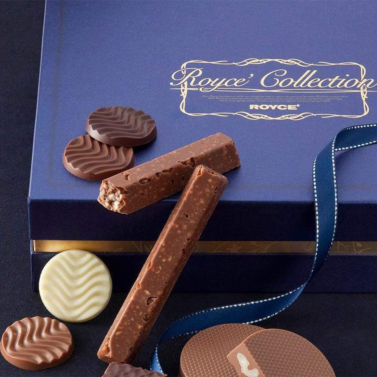 Open Royce’ Collection box displaying chocolate bars, round wafers in milk, dark, and white chocolate, with a branded dark blue gift box and ribbon.