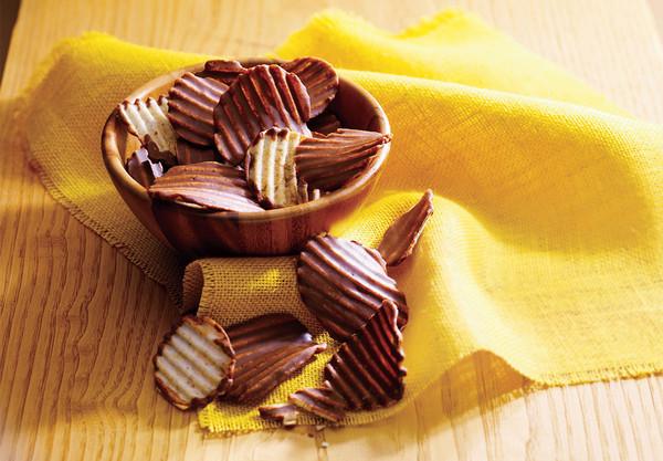 A small wooden bowl filled with chocolate-coated ridged potato chips, set on a wooden surface with a yellow cloth underneath.
